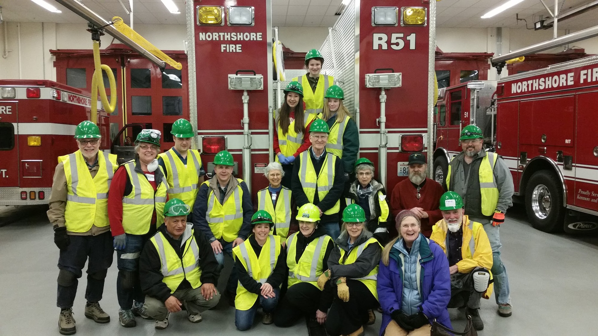 Community emergency response team members in safety vests and helmets pose in front of fire trucks during training.