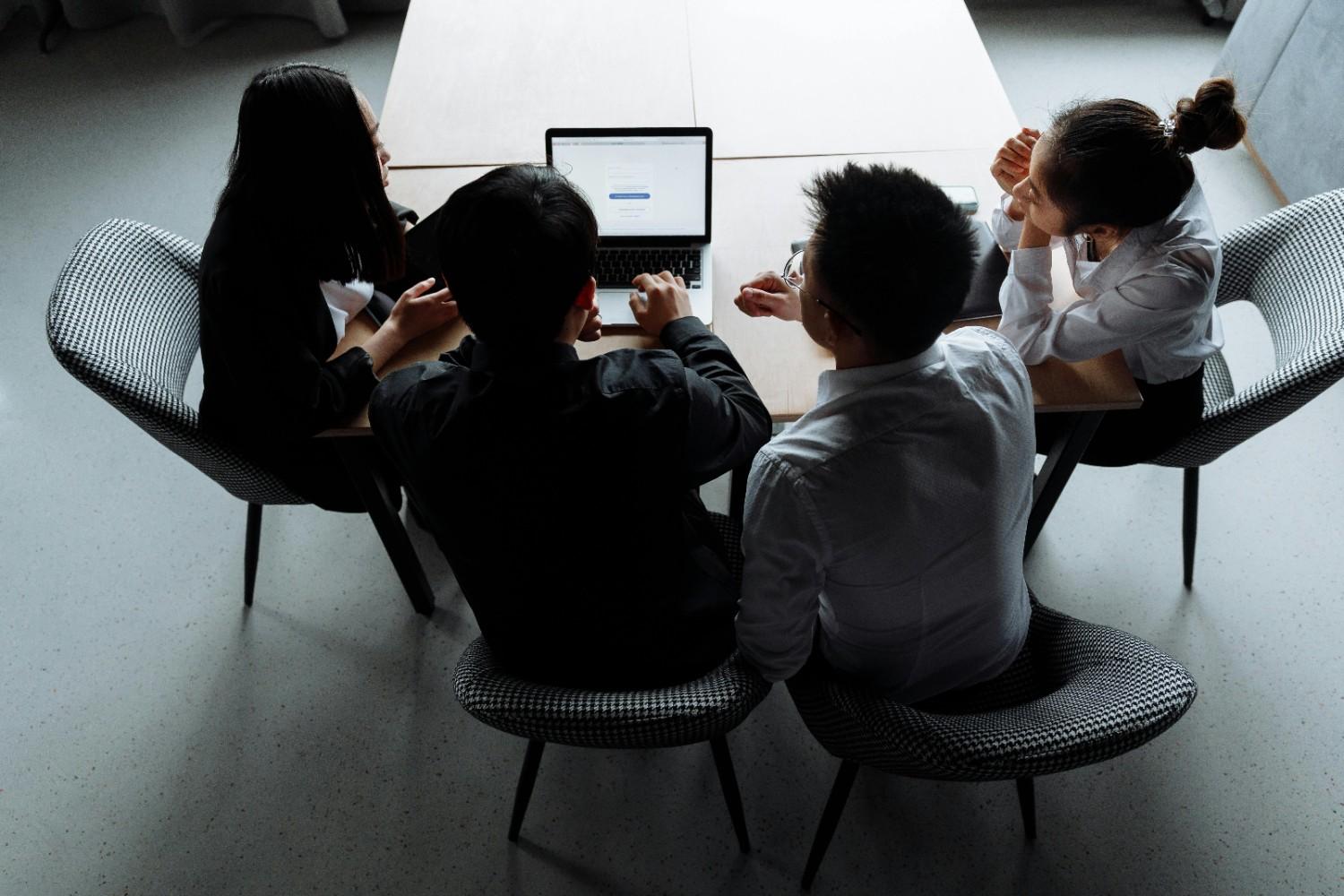 High-angle view of four HOA board members collaborating around a single laptop to manage community social media and digital communication updates.